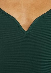Tissu vert à côtes avec un design à col en V profond. La texture semble lisse et légèrement brillante, mettant en valeur le décolleté structuré.