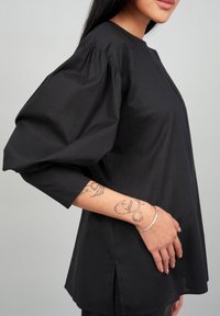 Black puff-sleeve blouse in lightweight fabric, featuring a round neckline, side slits, and a smooth texture. Silver bracelet on wrist.