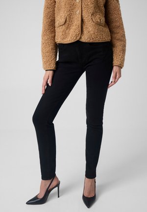 Frau trägt schwarze Slim-Fit-Hose, schwarze Slingback-Pumps mit hohem Absatz und eine beige strukturierte Jacke mit Knöpfen vor einfachem Hintergrund.