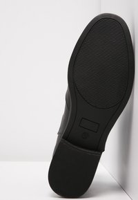 Suela de zapato negra con goma texturizada, presenta un borde superior suave y un pequeño acento rectangular en el lateral.