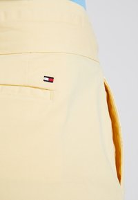 Detailní záběr na kapsu světle žlutých kalhot s malým obdélníkovým logem v červené, bílé a modré barvě vyšitým nad otvorem kapsy.