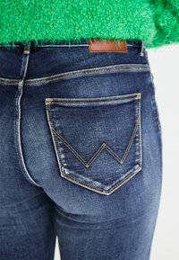 Tummansinisiä farkkuja, joissa on erottuva kulmikas takatasku desain, jossa on vastakkaista keltaista ommelta ja nahkaleima vyötäröllä.