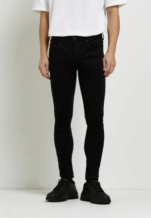 Person trägt schwarze, eng anliegende Jeans, schwarze Sneakers und ein weißes T-Shirt mit kurzen Ärmeln, steht vor einem einfarbigen hellen Hintergrund.