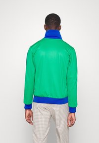 Vihreä takki sinisellä kauluksella ja kalvosimilla, jossa on sileä pinta ja joustava vyötärönauha. Päällä kuvioidut beiget housut.