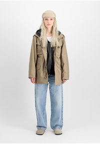 Khaki parka jacket over a black hoodie, light blue wide-leg jeans, and a beige cap. Features pockets and a drawstring at the waist.