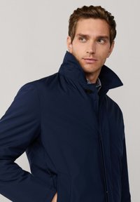 Man in een marineblauwe jas met verhoogde kraag, kijkend opzij, met zijn hand in de zak, tegen een effen lichte achtergrond.