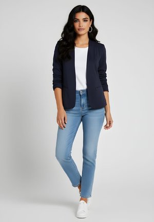 Mujer con cabello largo y oscuro, vestida con blazer azul marino, camiseta blanca, jeans azul claro y zapatillas blancas, de pie frente a un fondo liso.