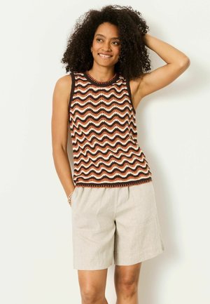 Frau mit lockigem Haar, die ein ärmelloses Oberteil mit wellenartigem Muster in Braun, Schwarz und Weiß sowie beige Shorts trägt, lächelnd mit einer Hand am Kopf.