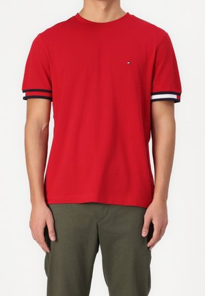 Man wearing bright red short-sleeve T-shirt with navy, white, and red trim on sleeves, paired with olive green pants, standing against white background.