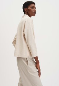 Top in maglia beige con vestibilità rilassata, scollatura rotonda e spalle scese. Presenta uno spacco sul retro ed è abbinato a pantaloni leggeri coordinati.