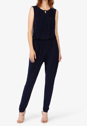 Kvinde iført en ærmeløs marineblå jumpsuit med samlet talje og nøglehulsudskæring, kombineret med nøgne højhælede sandaler.
