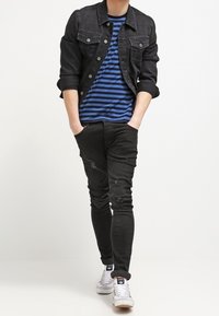 Ung man klädd i svart denimjacka, blå och svart randig skjorta, svarta rivna slim fit-jeans, vita sneakers, händerna i fickorna, går framåt.