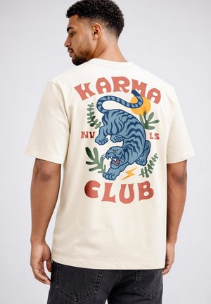 Man wearing cream t-shirt with colorful graphic of roaring tiger, plants, sun, and text "Karma Club" on the back.