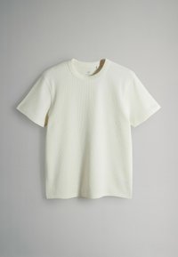 Camiseta blanca roto de manga corta con cuello redondo acanalado colgada frente a un fondo liso gris claro.