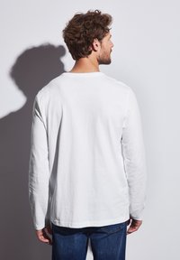 Camisa blanca de manga larga con cuello redondo, hecha de algodón, que presenta un ajuste relajado y una textura suave. Combinada con vaqueros de denim azul.