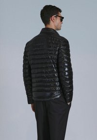 Schwarze, gesteppte Jacke mit glänzendem Finish. Verfügt über einen Kragen und eine taillierte Silhouette, kombiniert mit dunklen Hosen. Rückansicht, Modell trägt Sonnenbrille.
