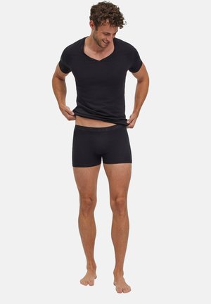 Mann trägt schwarzes V-Ausschnitt-T-Shirt und schwarze Boxershorts, schaut nach unten und hebt sein Shirt leicht an, barfuß auf weißem Hintergrund.