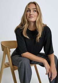 Black pleated top with wide sleeves, featuring a round neckline. Model sitting on a wooden chair, wearing gray tailored trousers.