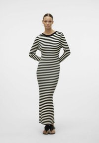 Robe maxi en tricot rayé noire et crème, avec manches longues, encolure ronde et silhouette ajustée. Portée avec des sandales noires à enfiler.