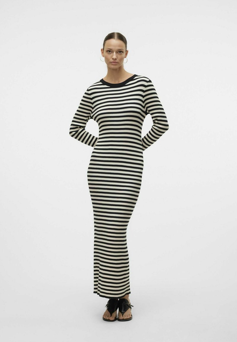 Robe maxi en tricot rayé noire et crème, avec manches longues, encolure ronde et silhouette ajustée. Portée avec des sandales noires à enfiler.