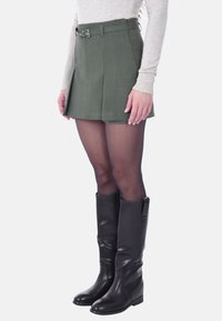 Mini gonna plissettata verde oliva con una cintura, realizzata in tessuto testurizzato, abbinata a stivali neri in pelle al ginocchio e calze trasparenti.