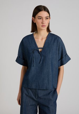 Kobieta ubrana w luźną, krótką ciemnoniebieską dżinsową bluzkę z wiązaniem z przodu oraz dopasowane szerokie spodnie, stojąca na tle jednolitego szarego tła.