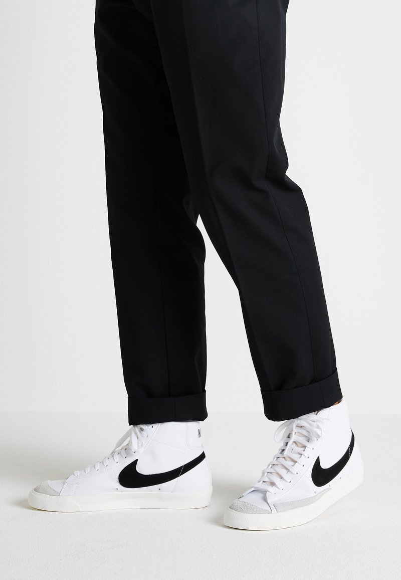 Baskets montantes blanches avec un swoosh Nike noir, un embout texturé et des accents en suède gris, accompagnées de pantalons noirs retroussés.