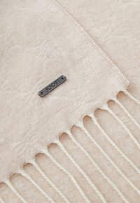 Lys beige skjerf laget av mykt materiale, med et subtilt mønster, ferdig med frynser, og et lite svart metallmerke som leser "JOOP!".