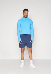 Ljusblå långärmad skjorta med halvdragkedja, i kombination med mörkblå shorts. Modellen bär vita sneakers med ankelsockor.