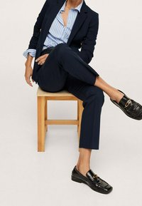 Granatowy garnitur z dopasowaną marynarką i wąskimi spodniami, zestawiony z jasnoniebieską koszulą w paski i czarnymi loafersami z złotym akcentem.