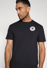 Must puuvillast lühikeste varrukatega T-särk, millel on ümar kaelus ja rindkere keskel valge ringikujuline logo, millel on tekst "Deutscher Fußball-Bund".