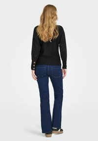 Maglione nero con lavorazione a maglia testurizzata e dettagli in bottoni ai polsini, abbinato a jeans a zampa d'elefante blu scuro e scarpe con piattaforma beige. Capelli lunghi e mossi.