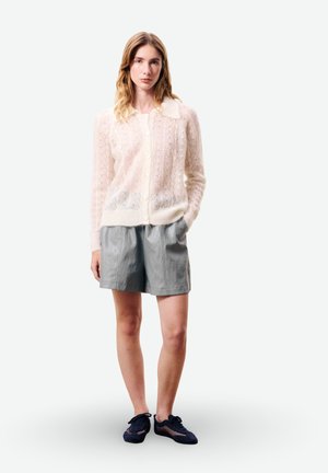 Femme portant un cardigan en maille crème transparent, un short taille haute gris et des chaussures noires à lacets, debout avec une main dans la poche.