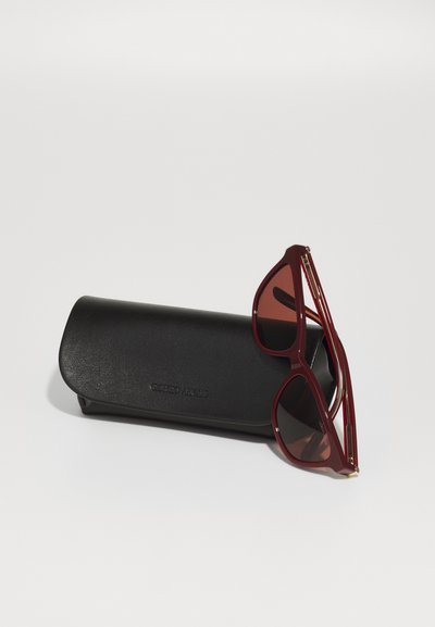 Ochelari de soare roșii cu rame geometrice și lentile degradate, așezați pe un capac texturat negru, cu branding embossat.