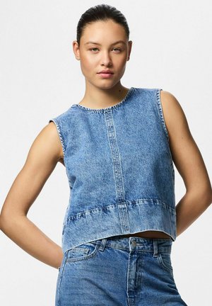 Noor naine tumedate juustega, kandes sinist lihvitud teksatoppi ilma varrukateta ja kõrge vöökohaga siniseid teksapükse, seisab ühe käega selja taga.