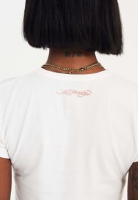 Witte katoenen t-shirt met korte mouwen, met roze geborduurde tekst op de bovenrug en gelaagde gouden kettingen.