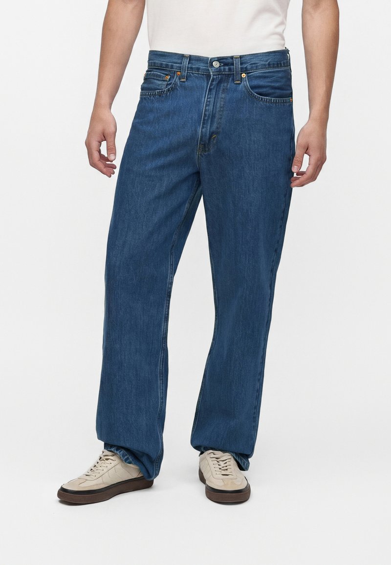 Mand iført løstsiddende blå denimjeans og beige sneakers, stående mod en ensfarvet hvid baggrund.