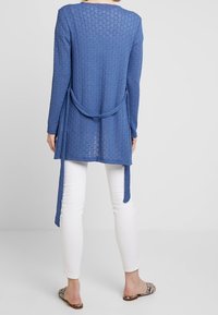 Cárdigan largo azul de manga larga hecho de tejido de punto texturizado, con un detalle de lazo en la cintura y un dobladillo asimétrico, combinado con pantalones blancos.