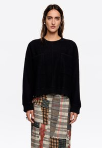 Suéter negro de punto con un corte holgado, que cuenta con dos bolsillos frontales y puños acanalados. Se lleva sobre una falda de cuadros con múltiples patrones.