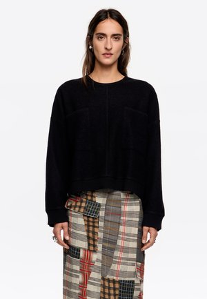 Suéter negro de punto con un corte holgado, que cuenta con dos bolsillos frontales y puños acanalados. Se lleva sobre una falda de cuadros con múltiples patrones.
