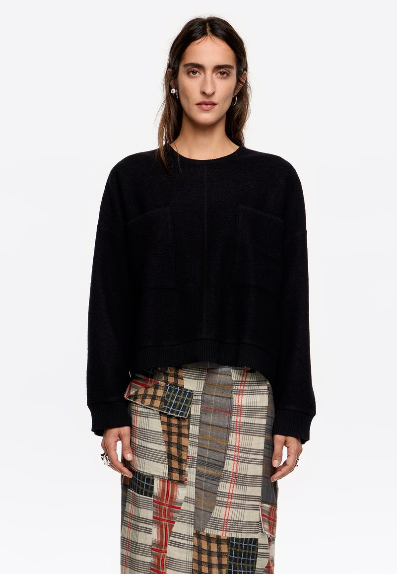 Suéter negro de punto con un corte holgado, que cuenta con dos bolsillos frontales y puños acanalados. Se lleva sobre una falda de cuadros con múltiples patrones.