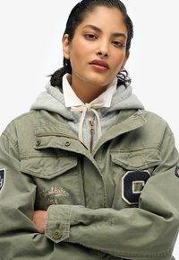 Oliwkowa kurtka robocza z wieloma kieszeniami, haftowanymi detalami i ciemnoniebieską łatą. Nałożona na szary hoodie z sznurkiem.