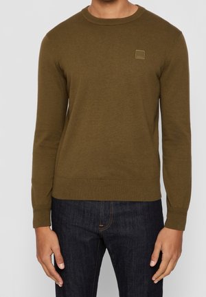 Mann trägt olivegrünen langärmligen Strickpullover mit rundem Ausschnitt und kleinem quadratischem Logo auf der Brust, kombiniert mit dunkelblauen Jeans.