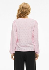 Woman with wavy blonde hair wearing a long-sleeve light pink eyelet blouse and black pants, viewed from the back on a white background.