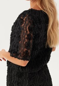 Sort kjole med et tekstureret blomsterdesign, med gennemsigtige korte ærmer og en tætsiddende overdel. Stof ser blødt ud med lagdelte udsmykninger.