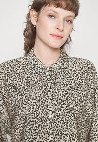 Camisa bege de botão com padrão manchado a preto, colarinho e bolsos frontais, feita de um tecido leve e texturizado.