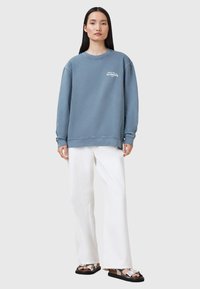 Modrá mikina s kulatým výstřihem a dlouhými rukávy, s malým logem na hrudi, doplněná o bílé kalhoty s širokými nohavicemi a sandály.