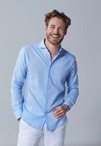 Lichtblauwe langetermijnhemd van katoen, met een button-down kraag, knoopsluiting aan de voorkant en een afgeronde onderkant, gedragen met witte broek.