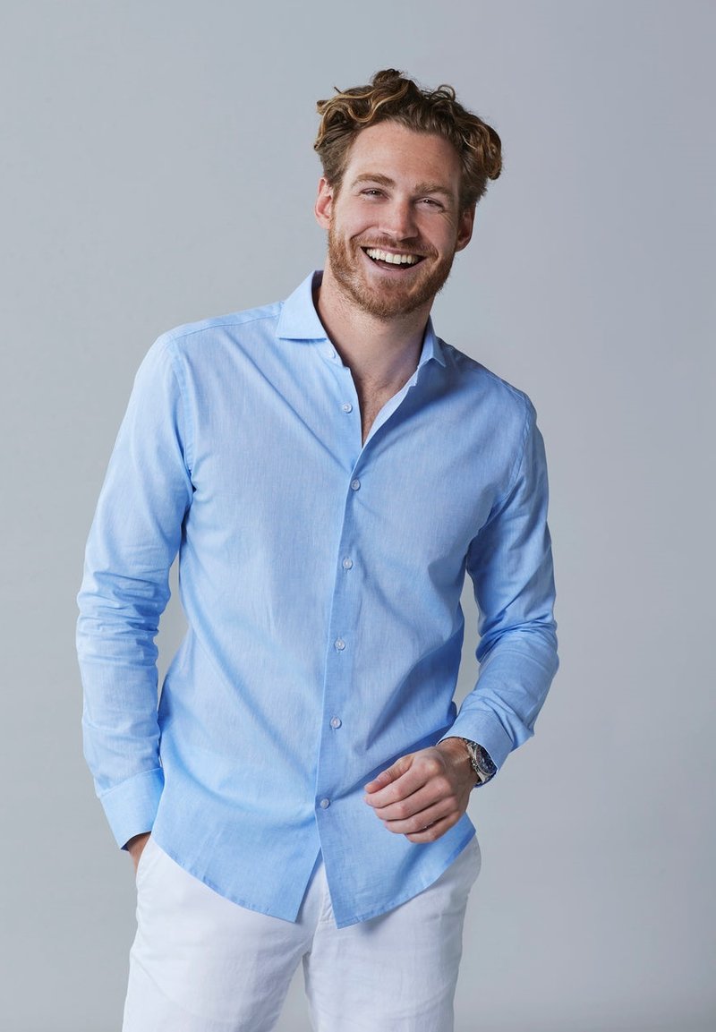 Lichtblauwe langetermijnhemd van katoen, met een button-down kraag, knoopsluiting aan de voorkant en een afgeronde onderkant, gedragen met witte broek.