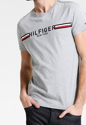 Lichtgrijs T-shirt met korte mouwen met de tekst "HILFIGER NEW YORK" en rode, witte en zwarte strepen, gedragen door een man met zijn handen in de zakken.